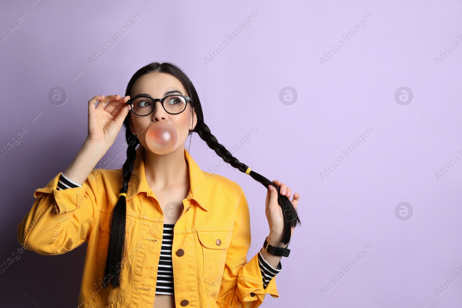 Photo of Fashionable young woman with braids blowing bubblegum on lilac background, space for text