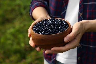 Photo of Woman holding wooden bowl of bilberries outdoors, closeup. Space for text