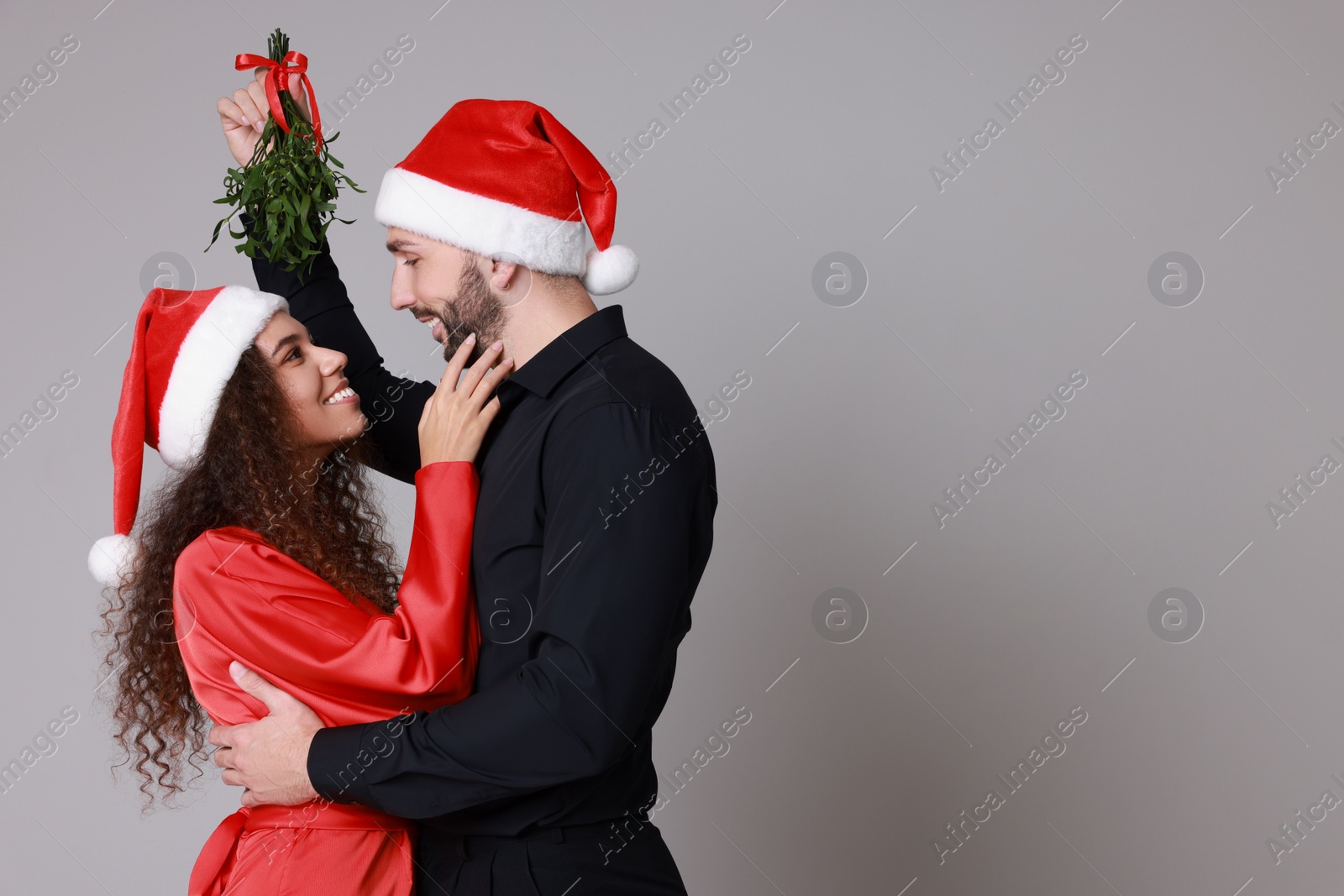 Photo of Lovely couple in Santa hats under mistletoe bunch on grey background. Space for text