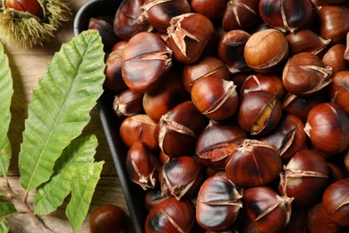 Photo of Delicious roasted edible chestnuts and green leaves on wooden table, top view