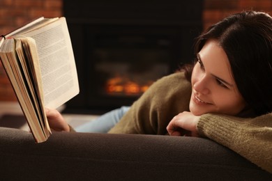 Photo of Young woman reading book on sofa near fireplace at home