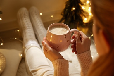 Photo of Woman holding cup with hot drink near festive lights at home, closeup