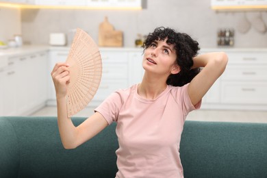 Photo of Young woman waving hand fan to cool herself on sofa at home