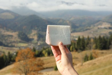 Woman holding piece of delicious cheese against mountain landscape, closeup