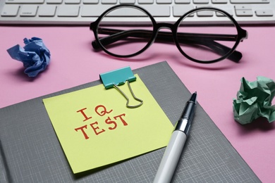 Photo of Sticker note with text IQ Test, notebook, pen, crumpled paper balls, keyboard and glasses on pink background, closeup