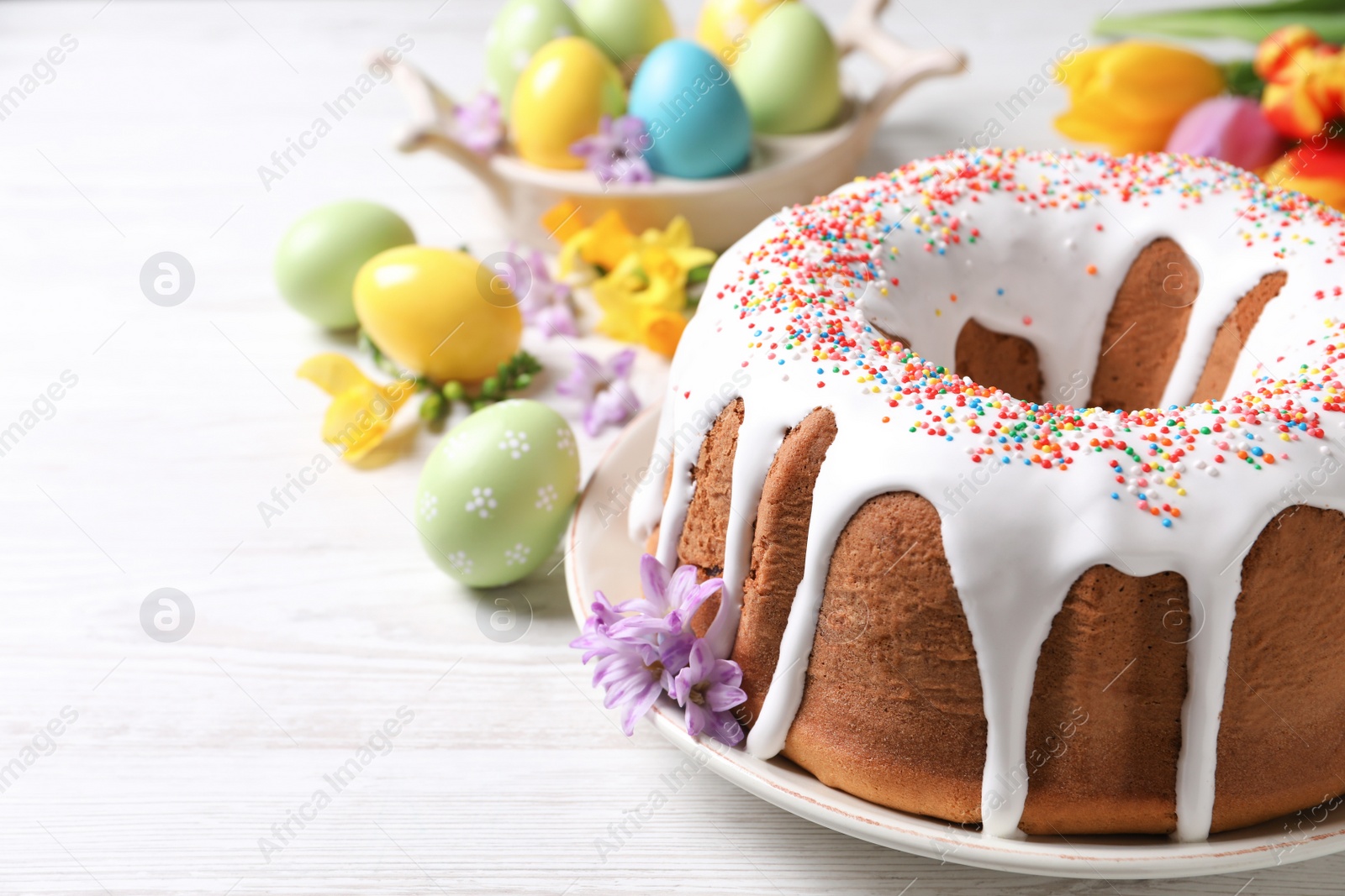 Photo of Glazed Easter cake with sprinkles, painted eggs and flowers on white wooden table. Space for text