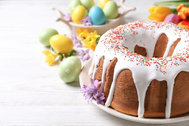 Photo of Glazed Easter cake with sprinkles, painted eggs and flowers on white wooden table. Space for text