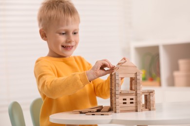 Cute little boy playing with wooden house at white table indoors. Child's toy