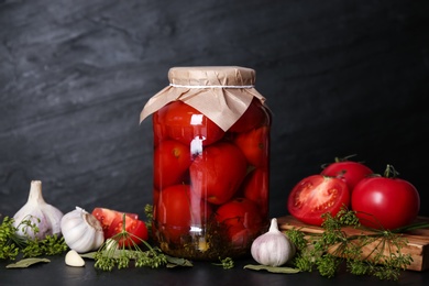 Glass jar of pickled tomatoes and ingredients on black table