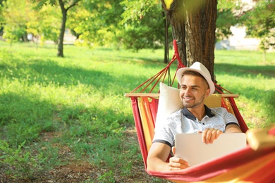 Photo of Young man with laptop resting in comfortable hammock at green garden
