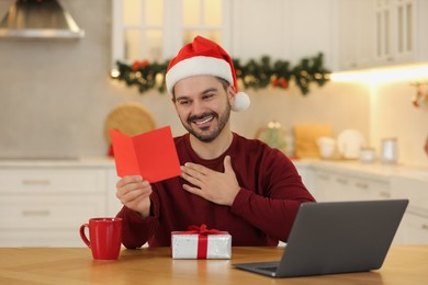 Photo of Celebrating Christmas online with exchanged by mail presents. Happy man reading greeting card during video call on laptop at home
