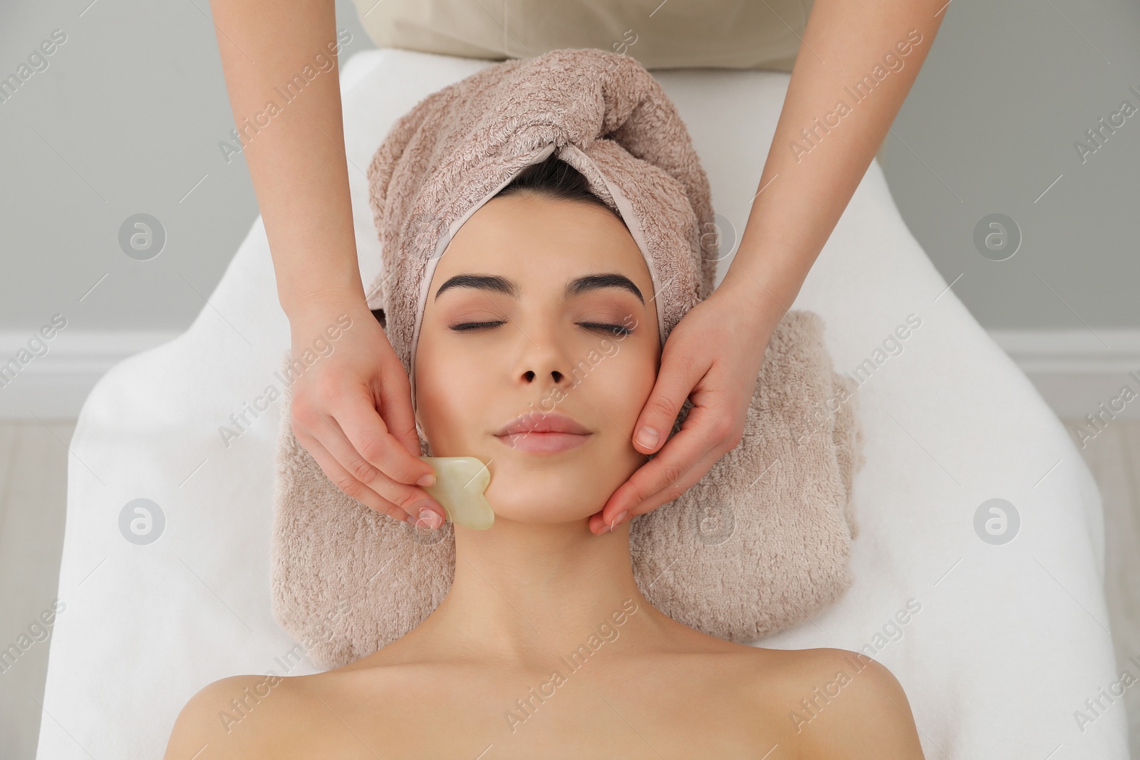 Photo of Young woman receiving facial massage with gua sha tool in beauty salon, top view