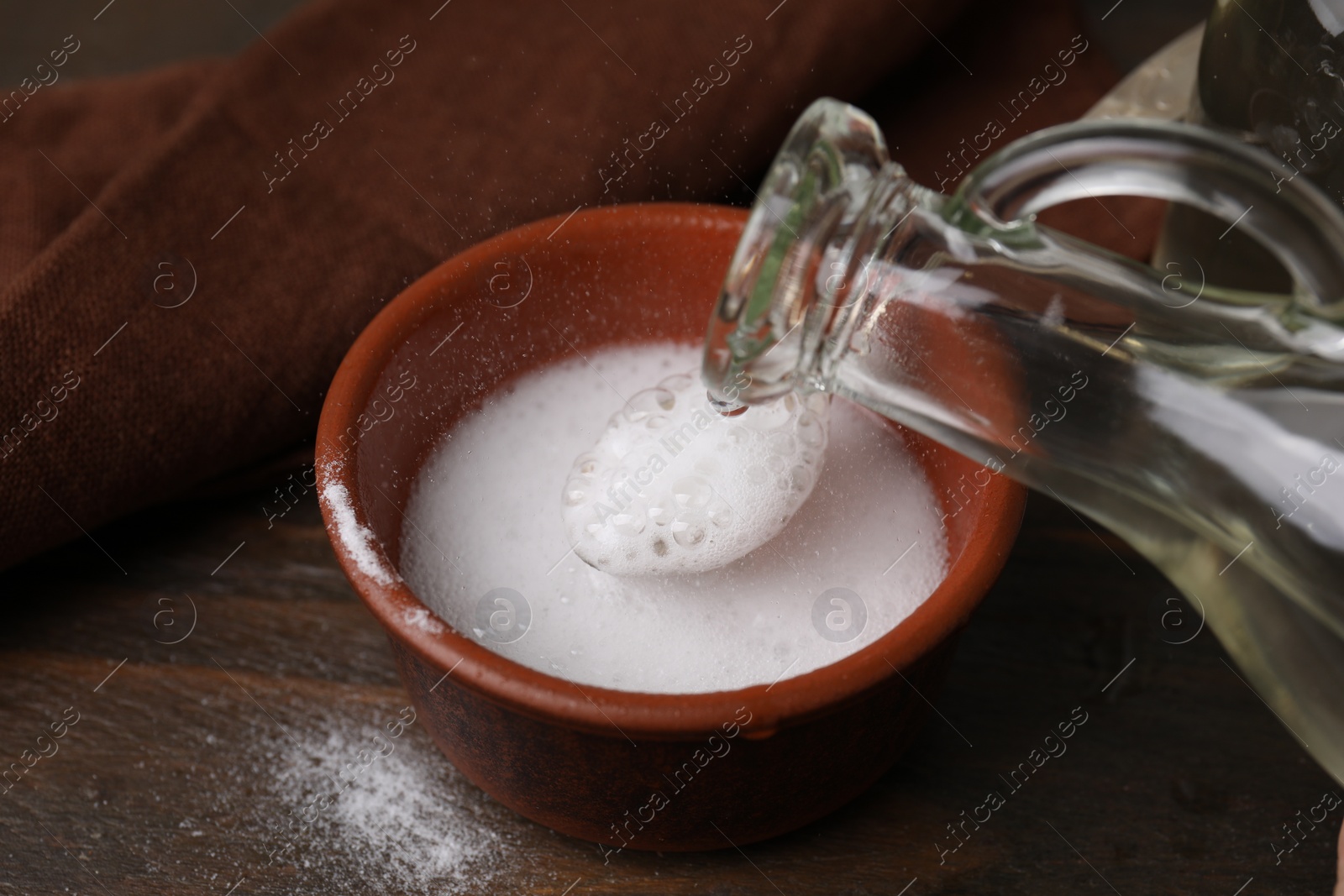 Photo of Pouring vinegar into spoon with baking soda over bowl at wooden table, closeup