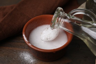 Pouring vinegar into spoon with baking soda over bowl at wooden table, closeup