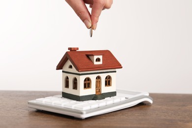 Mortgage concept. Woman putting coin into house model at wooden table against white background, closeup