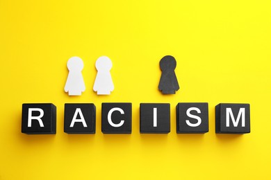 Photo of Word Racism made of cubes near black and white human paper figures on yellow background, flat lay