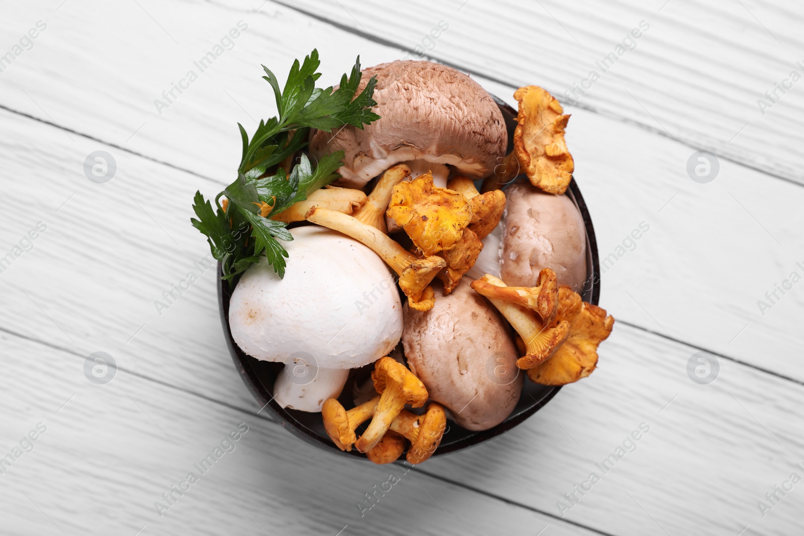 Photo of Bowl of different mushrooms and parsley on white wooden table, top view