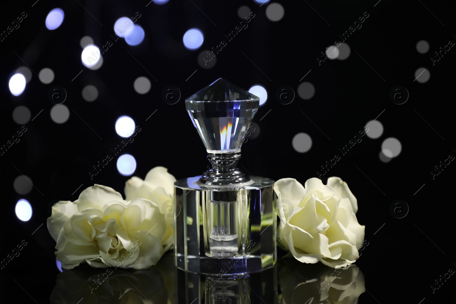 Photo of Perfume and flowers against blurred lights on black background, closeup