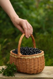 Photo of Woman taking wicker basket with bilberries at wooden table outdoors, closeup