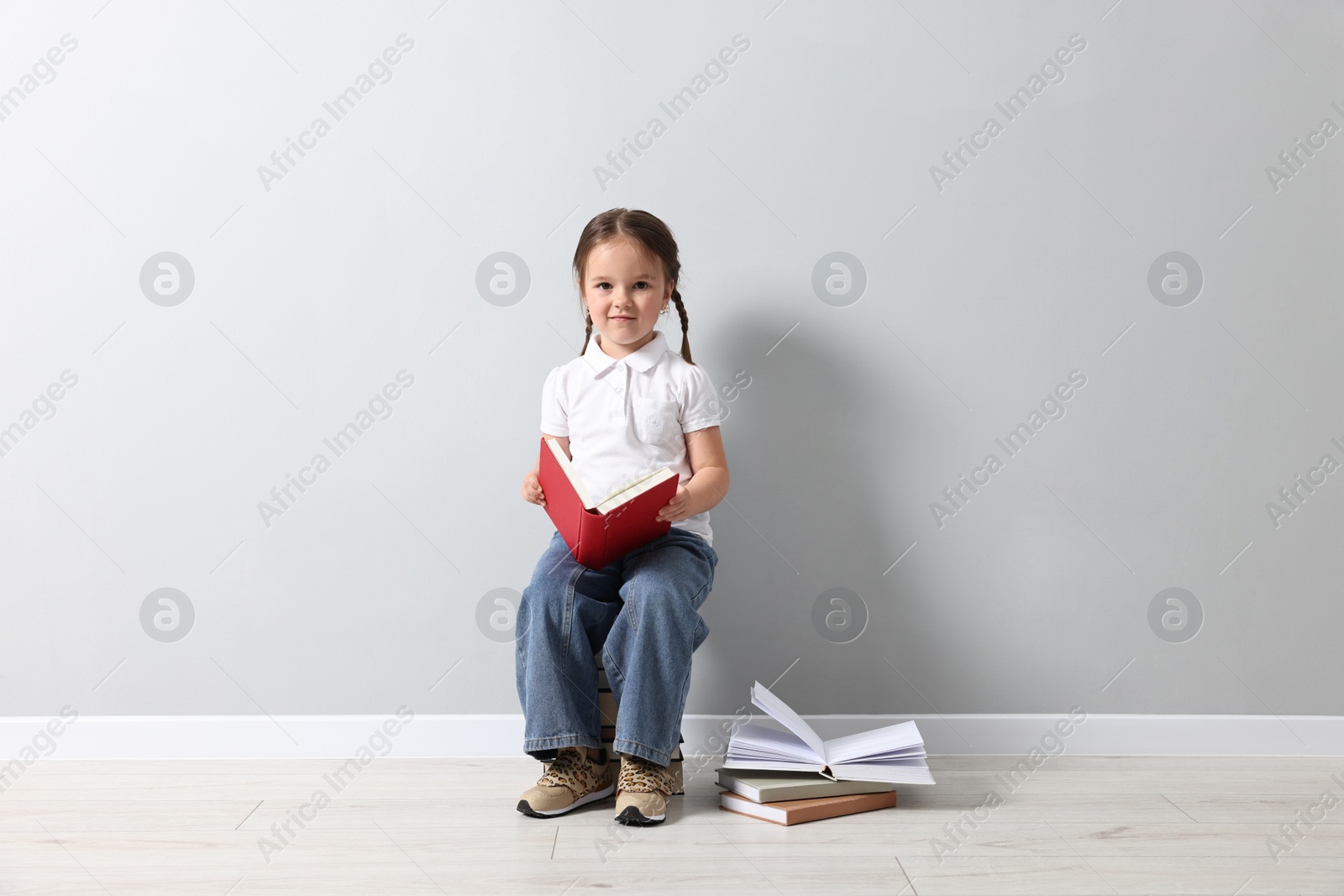 Photo of Cute little girl sitting on stack of books near light grey wall