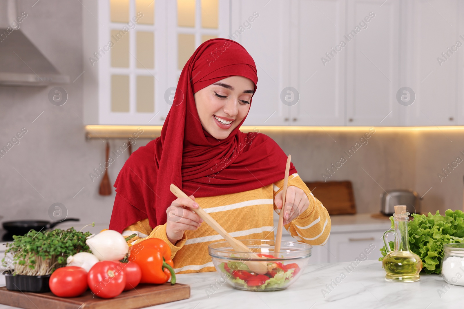 Photo of Muslim woman making delicious salad with vegetables at white table in kitchen