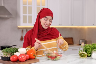 Muslim woman making delicious salad with vegetables at white table in kitchen