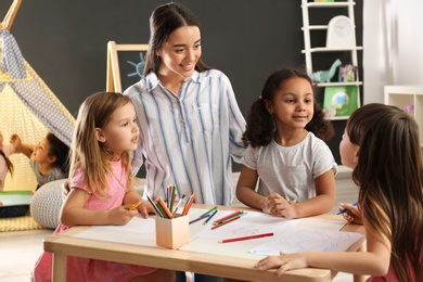 Photo of Cute little children with nursery teacher drawing at table in kindergarten. Indoor activity