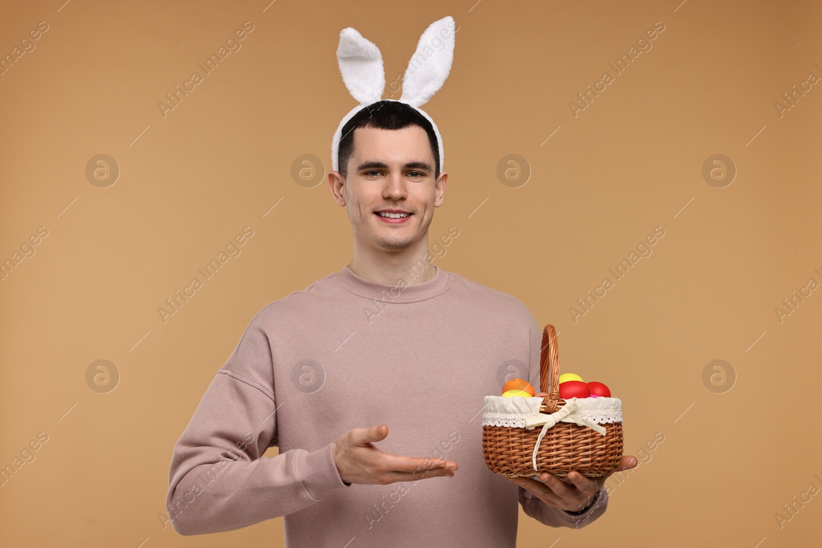 Photo of Easter celebration. Handsome young man with bunny ears holding basket of painted eggs on beige background