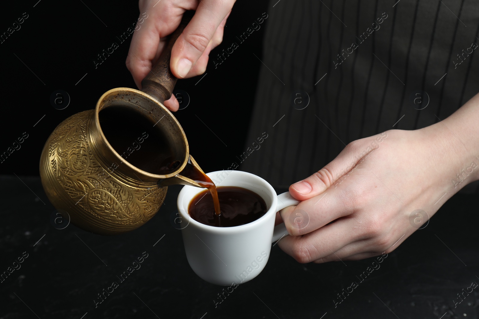 Photo of Turkish coffee. Woman pouring brewed beverage from cezve into cup at black table, closeup