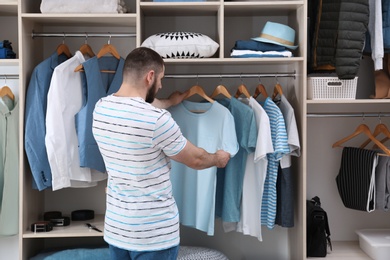 Photo of Man choosing outfit from large wardrobe closet with stylish clothes and home stuff