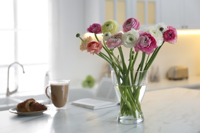 Photo of Beautiful fresh ranunculus flowers near cup of coffee, croissant and book on table in kitchen