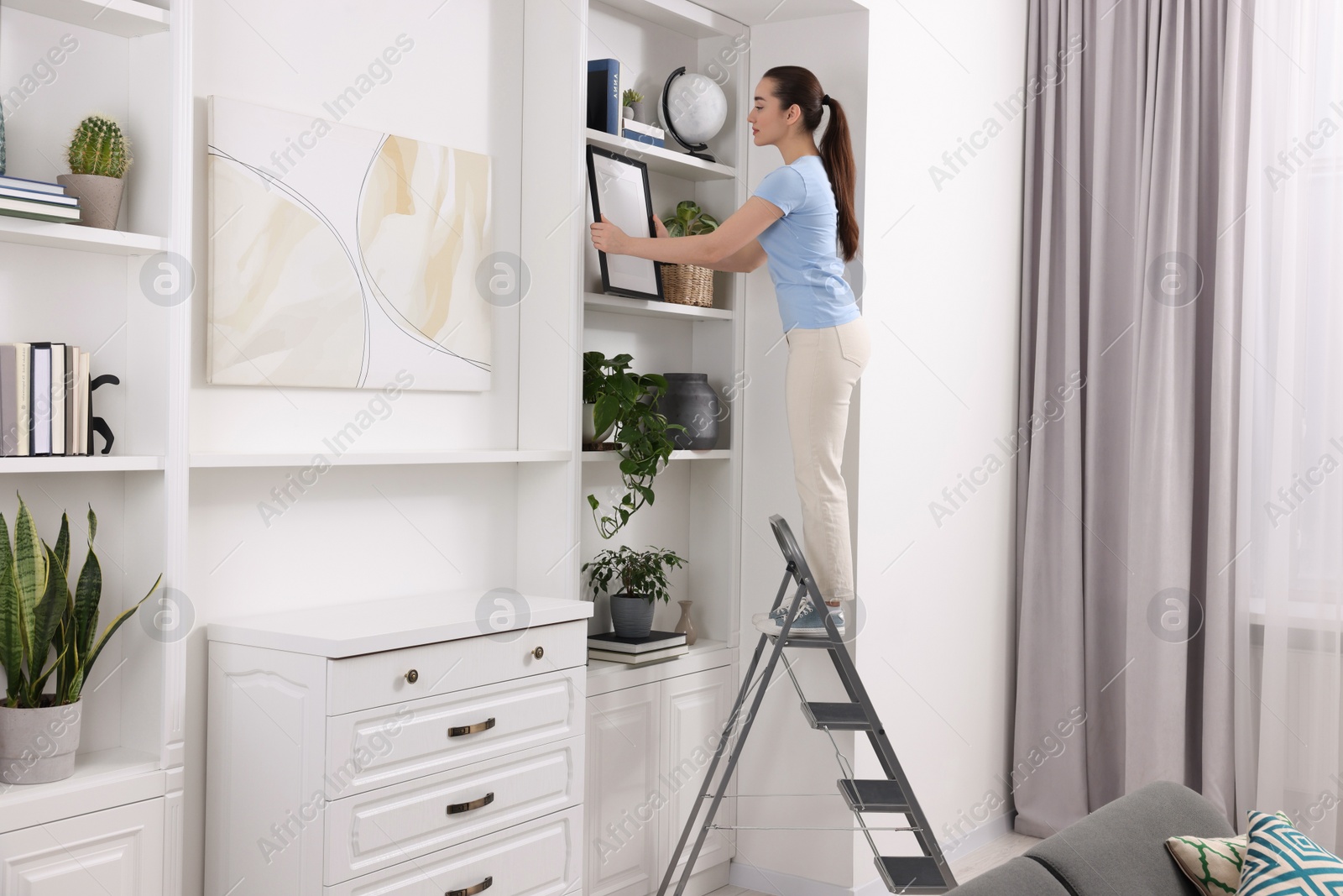 Photo of Woman on ladder with picture near shelves at home