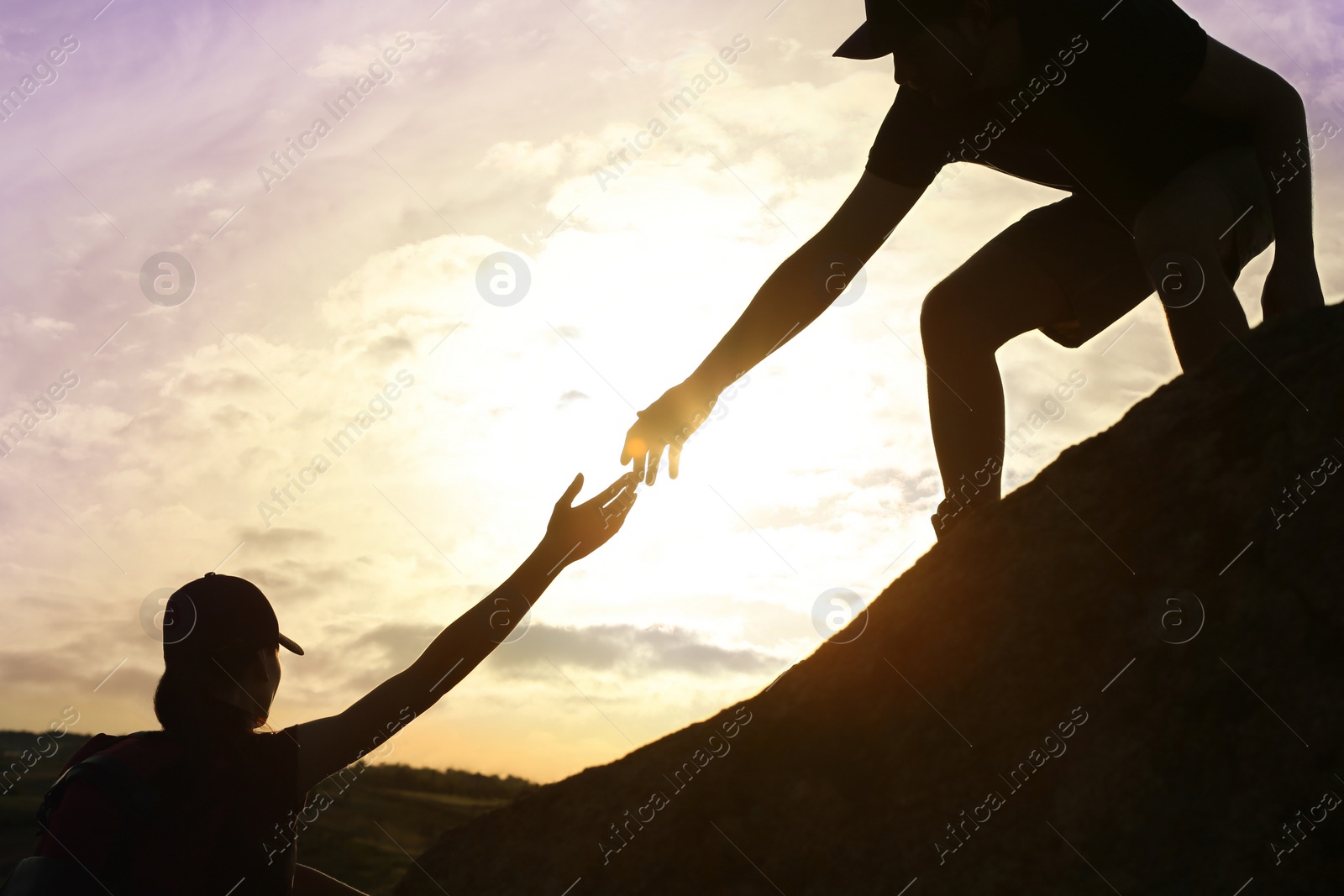 Photo of Silhouettes of man and woman helping each other to climb on hill against sunset