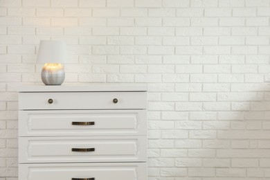 Modern chest of drawers with lamp near white brick wall. Space for text