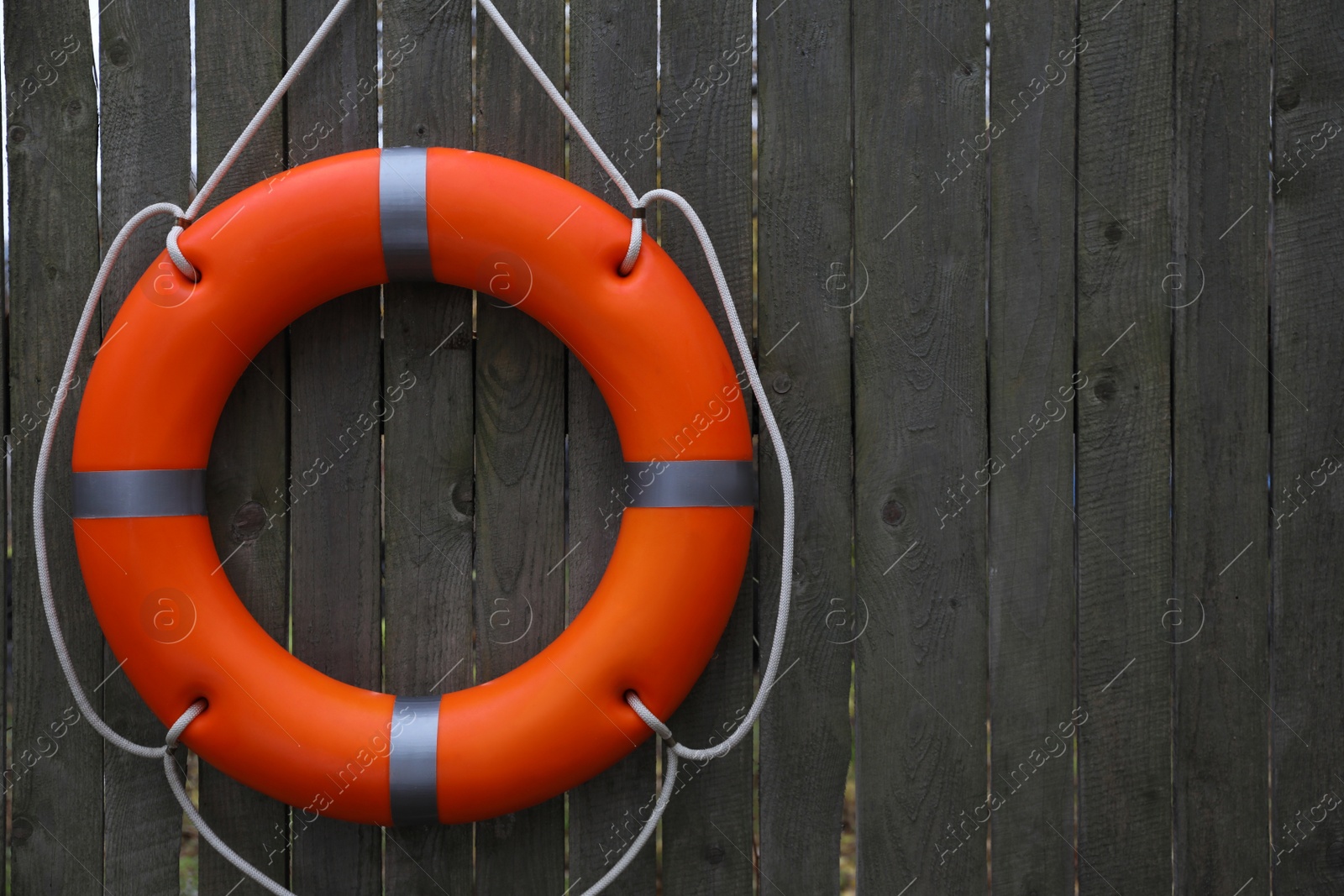 Photo of Orange lifebuoy hanging on grey wooden fence, space for text. Rescue equipment