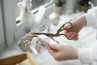 Photo of Woman cutting ribbon near Christmas baubles on window sill indoors, closeup