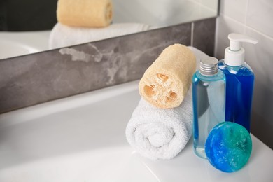 Photo of Loofah sponge, rolled towel and cosmetic products on sink in bathroom, space for text