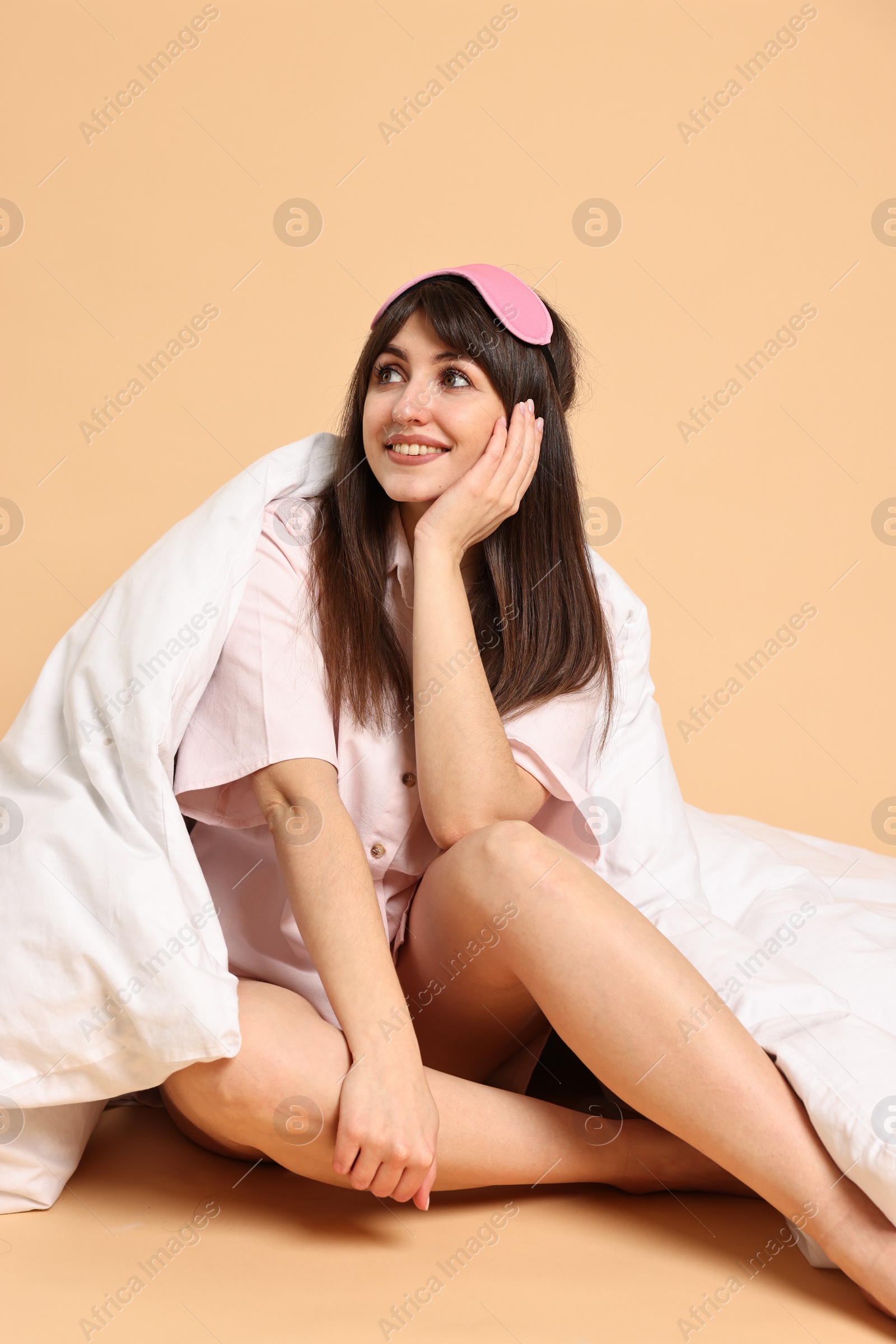 Photo of Happy woman with pyjama and sleep mask wrapped in blanket on beige background