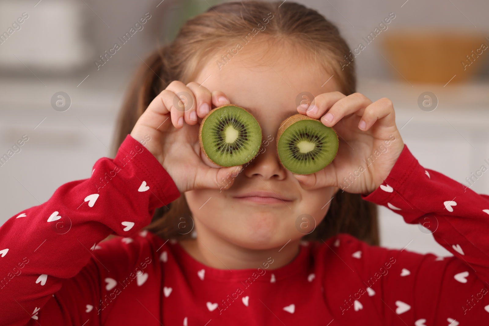 Photo of Funny girl covering eyes with halves of fresh kiwi indoors
