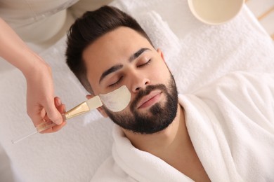 Photo of Cosmetologist applying mask on man's face in spa salon, top view