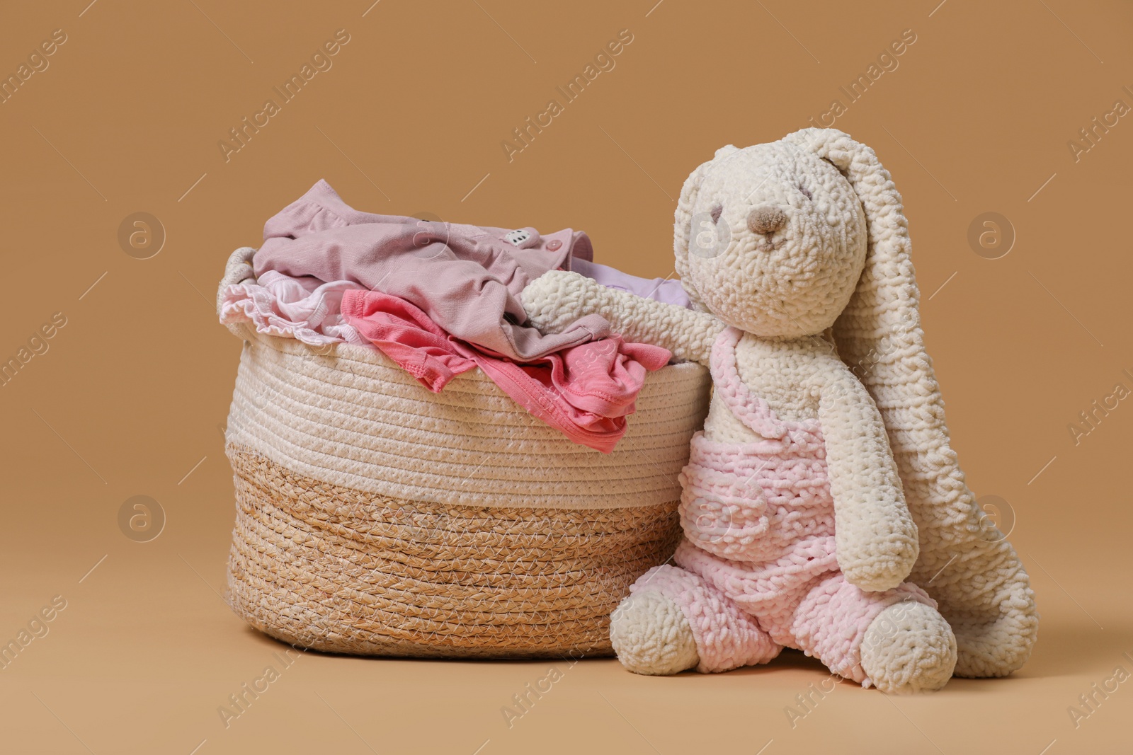 Photo of Laundry basket with baby clothes near soft toy on light brown background
