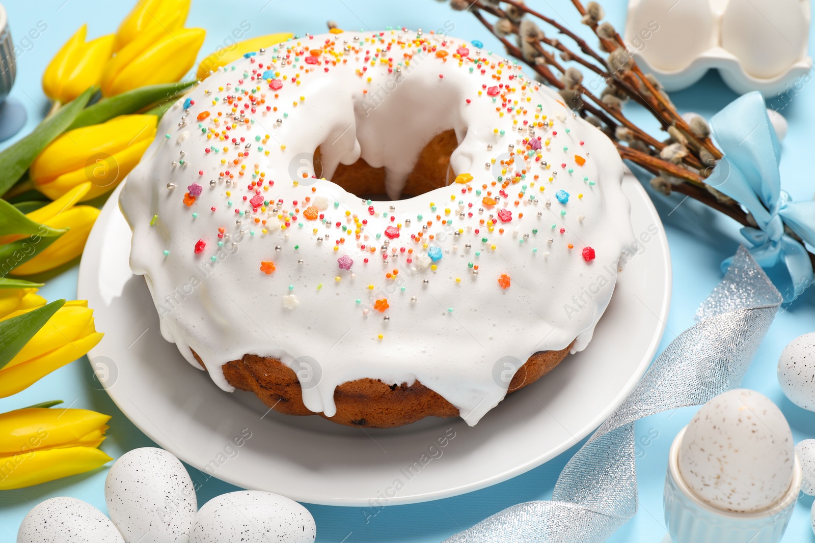 Photo of Delicious Easter cake decorated with sprinkles near beautiful tulips, willow branches and painted eggs on light blue background