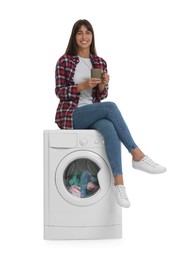 Photo of Beautiful woman with cup of drink sitting on washing machine against white background