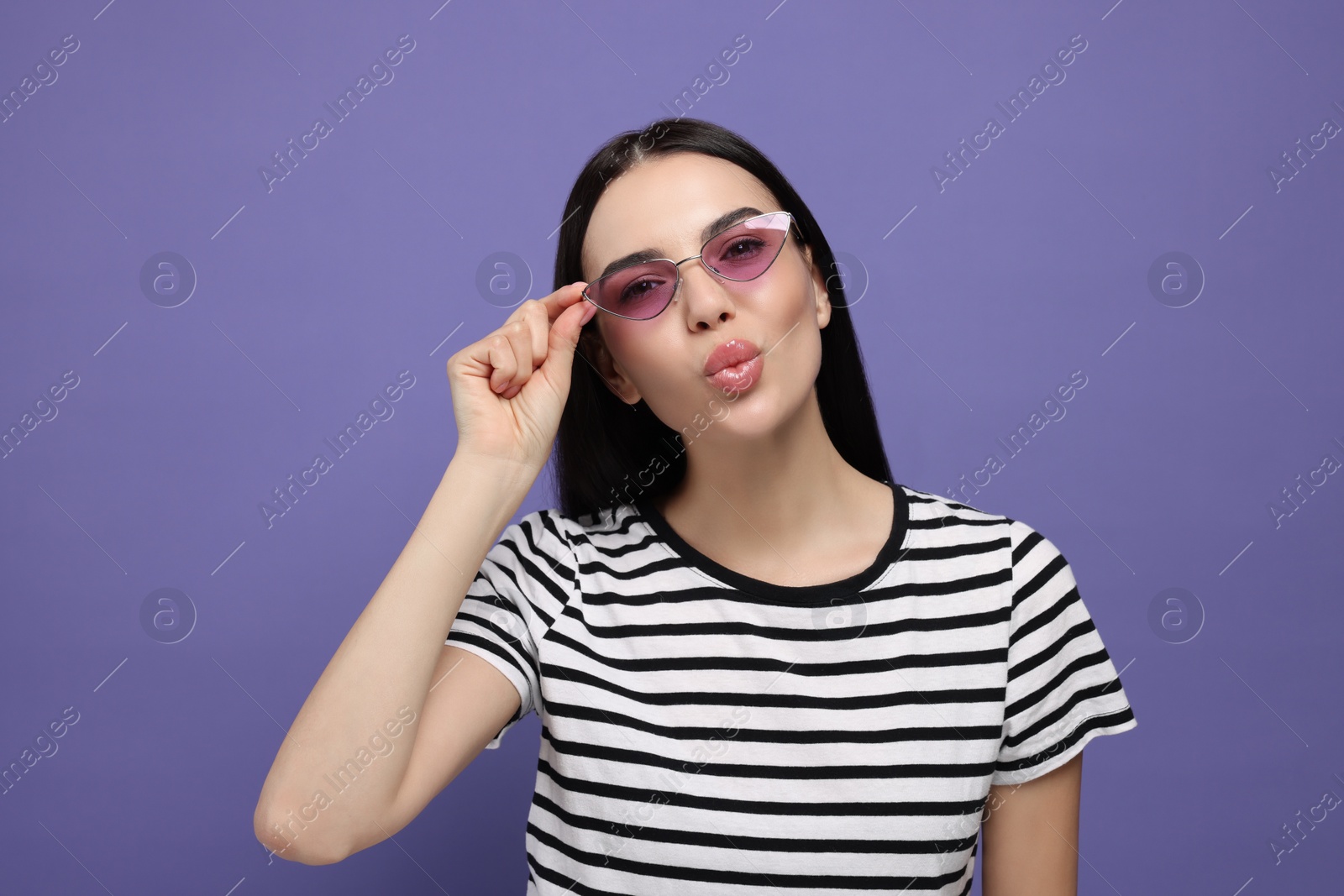 Photo of Beautiful young woman in stylish sunglasses blowing kiss on purple background