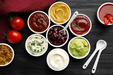 Photo of Different tasty sauces in bowls, tomatoes and spoons on black wooden table, flat lay