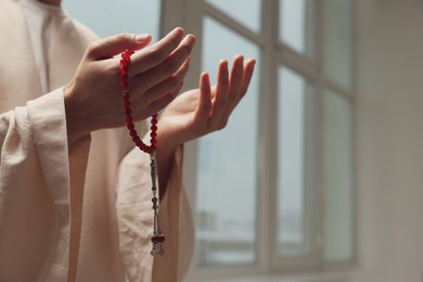 Photo of Muslim man with misbaha praying near window indoors, closeup. Space for text