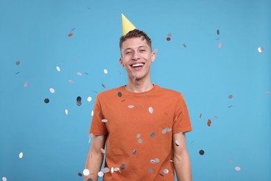 Photo of Happy man in party hat under falling confetti on light blue background