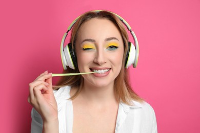 Fashionable young woman with bright makeup and headphones chewing bubblegum on pink background