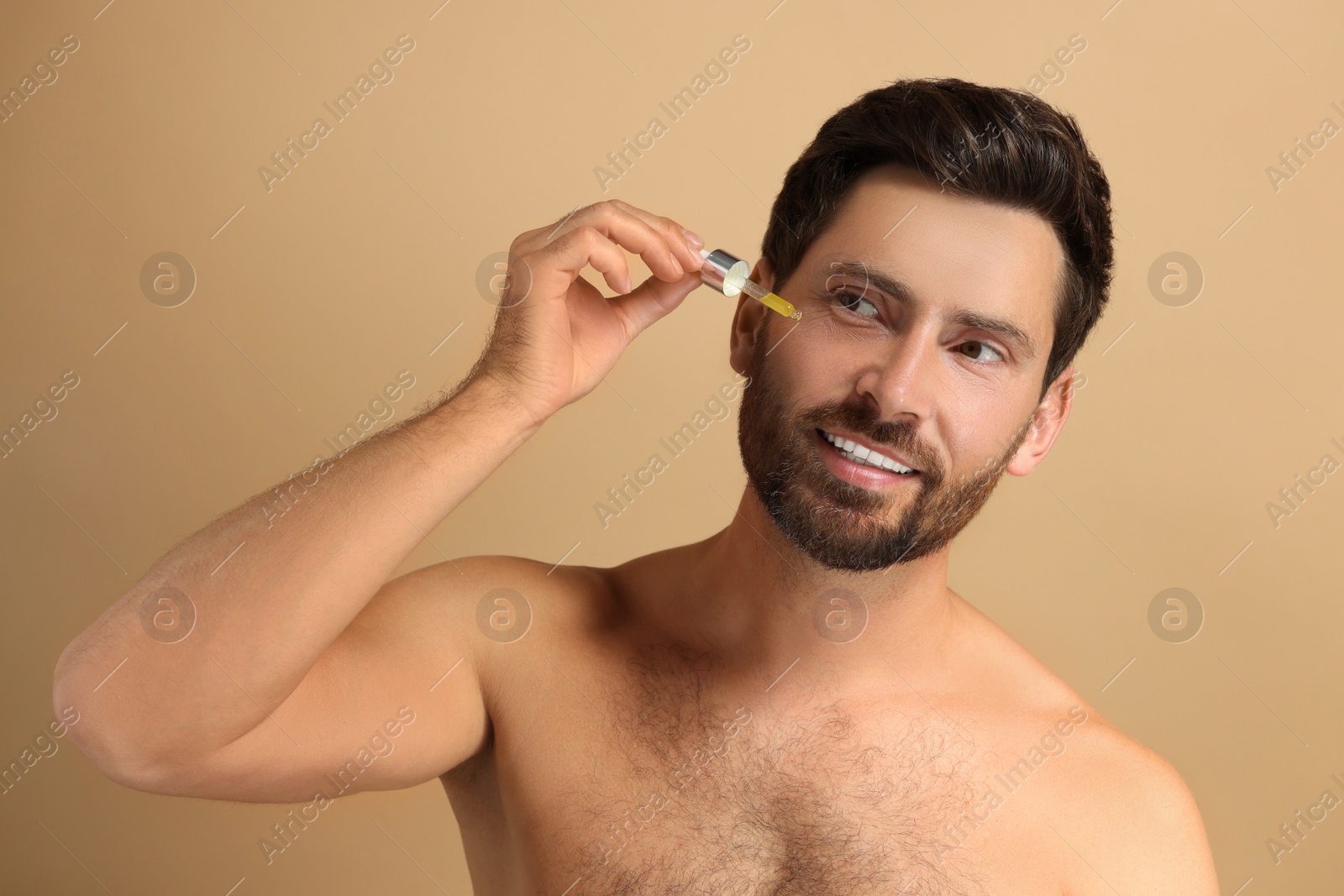 Photo of Smiling man applying cosmetic serum onto his face on beige background