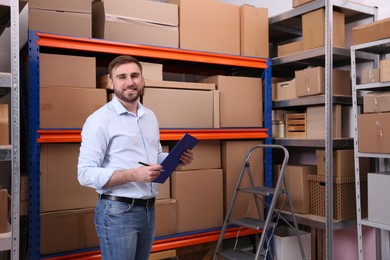 Photo of Young businessman with clipboard near rack of cardboard boxes at warehouse